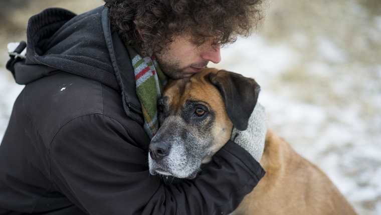 Young Caucasian man wearing a winter coat and a scarf hugging his pet dog lovingly to comfort him as they are out for a walk through a snowy forest during winter. The dog is a mixed breed and brown and black in color with a sad expression on his face.