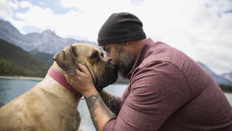 Affectionate bearded man with tattoos kissing dog at remote lakeside