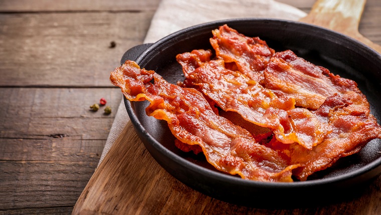 Hot fried crispy bacon slices in skillet on wooden background.