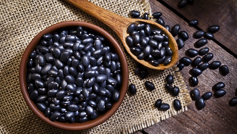 Top view of a brown bowl filled with black beans shot on rustic wood table. A wooden spoon is beside the bowl with some beans on it. DSRL studio photo taken with Canon EOS 5D Mk II and Canon EF 100mm f/2.8L Macro IS USM