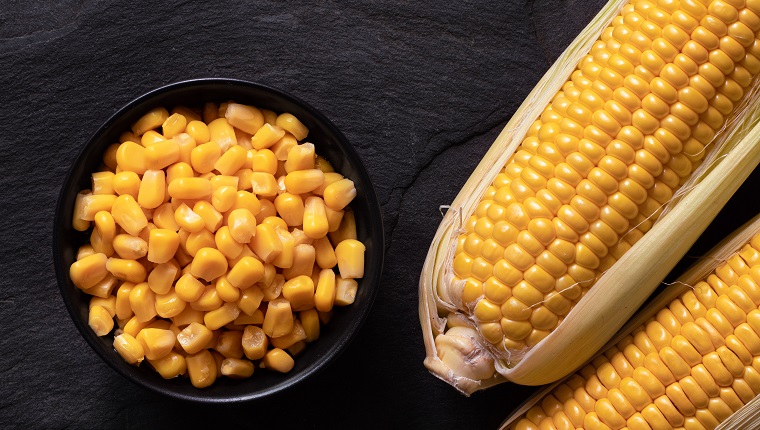 sweet corn in a black ceramic bowl next to two corn cobs in husks on black slate. Top view.