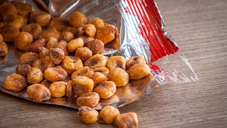 "Corn Snack Spilling from an Open Bag, CloseupA disposable bag sits on a wooden table, torn wide open, its silvery lining and its corn nuts in plain view."