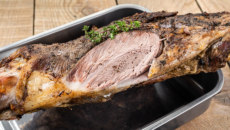 Whole oven roasted goat leg meat with thyme in a steel tray. Wooden background. Top view.