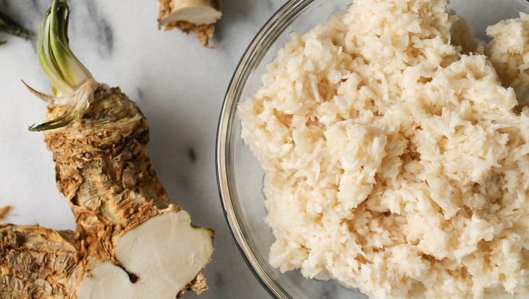 An overhead, extreme close up horizontal photograph of some homegrown horseradish roots and a glass bowl of prepared horseradish on a marble countertop.