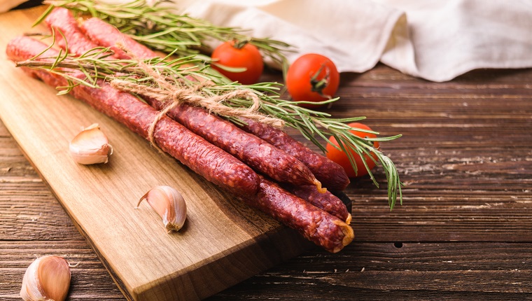 Kabanosy - smoked traditional polish pork sausages on wooden cutting board. With rosemary herb, garlic cloves and tomatoes. On rustic wooden table