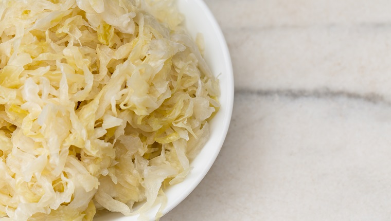 An overhead close up horizontal photograph of a white bowl sitting on a marble counter and containing some fresh, homemade sauerkraut.