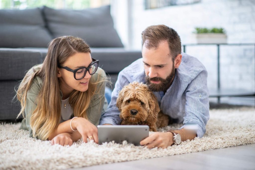 A young couple and their pet lay on the floor of their living room with a tablet out in front of them, as they tackle costs of owning a dog.